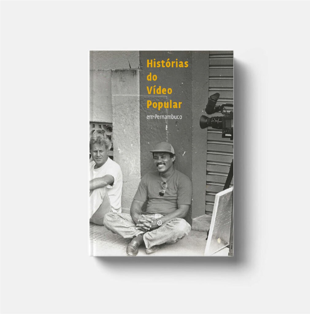 Imagem da capa do livro Histórias do Vídeo Popular em Pernambuco. Título escrito nas cores amarelo e branco sobre foto de dois homens sentados numa calçada descansando e uma câmera de vídeo do lado direito da imagem em preto e branco